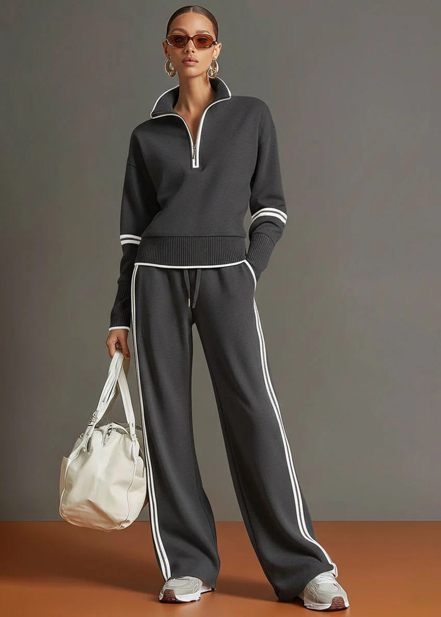 Woman wearing a dark gray tracksuit with white stripes, holding a white bag against a gray background.